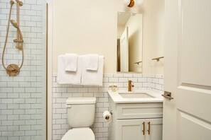 Ein Badezimmer mit weiß gefliesten Wänden, einer Toilette, einem Waschbecken mit Spiegel darüber und einer Dusche mit goldfarbener Duschbrause.