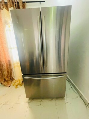 A stainless steel refrigerator in a kitchen with a tiled floor and a curtain. - Peaceful 4-bedroom house with AC in charming Old Yundum (Old yundum)