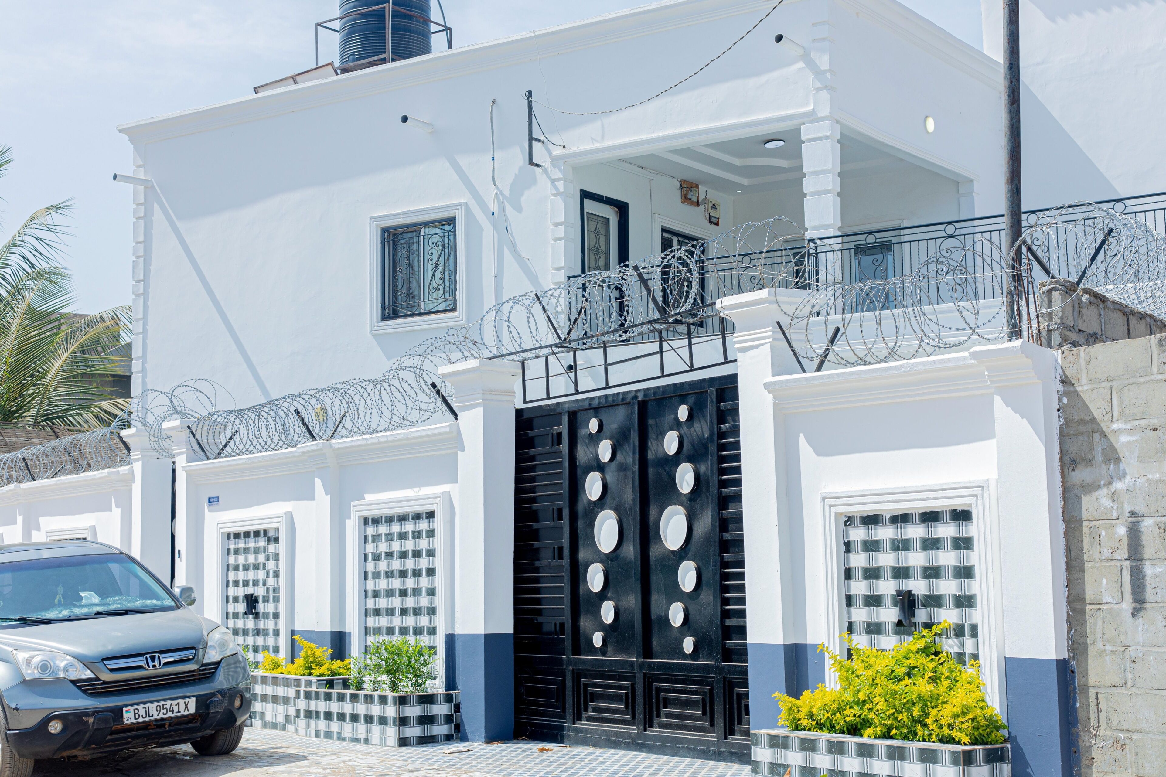 A white building with a black gate, a balcony with barbed wire, and a car parked in front.