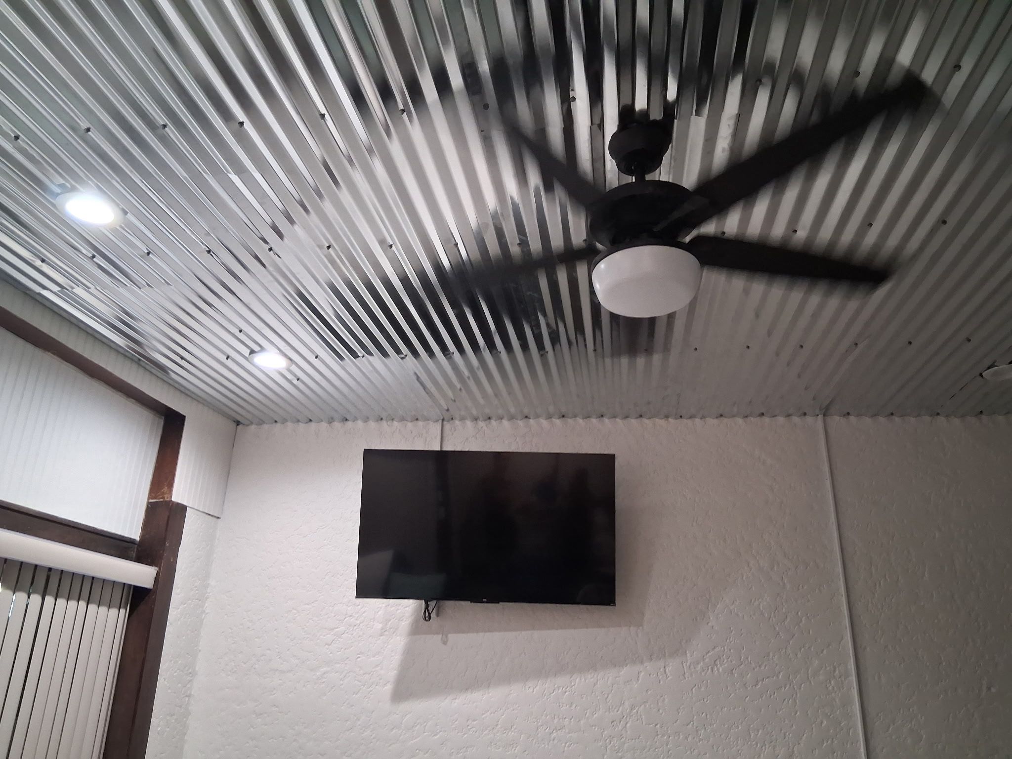 A room with a ceiling fan, a mounted flat-screen TV, and a textured white wall.