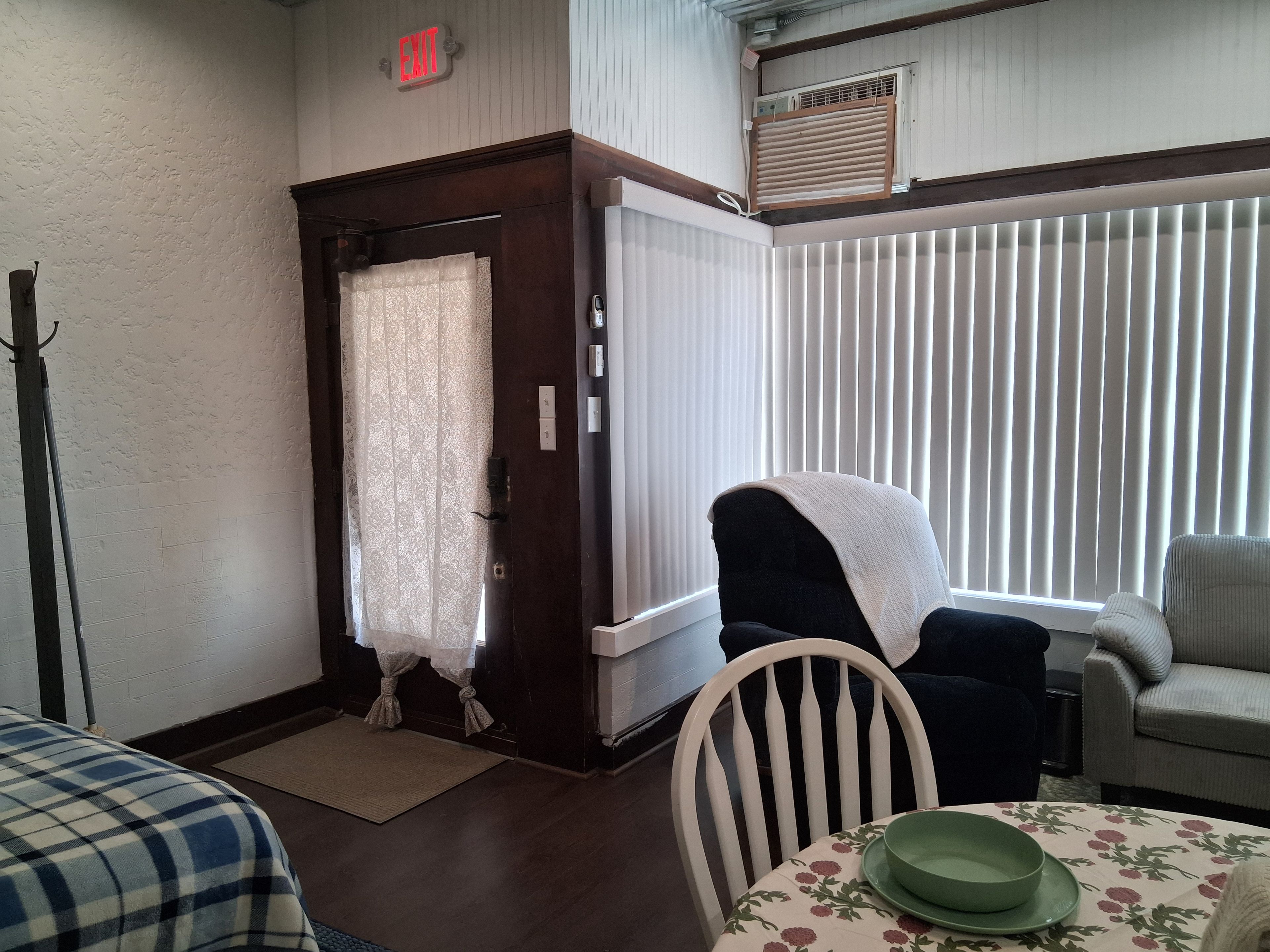 Ein Zimmer mit Tür, einem Sessel, einem Tisch mit Blumenmuster und einem Fenster mit Jalousien.
