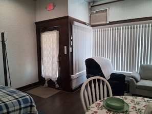 A room with a door, a chair, a table with a floral tablecloth, and a window with blinds.