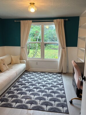 Ein Zimmer mit einer Couch, einem gemusterten Teppich, einem Schreibtisch mit Stuhl und einem Fenster mit Vorhängen.