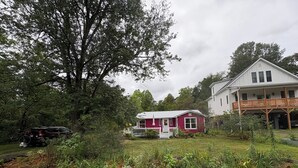 A house with a white exterior and a red front, surrounded by a garden with various plants and a large tree. - Peaceful Vintage Charm in North Asheville (Asheville)