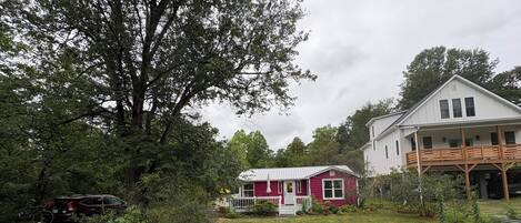 A house with a white exterior and a red front, surrounded by a garden with various plants and a large tree.