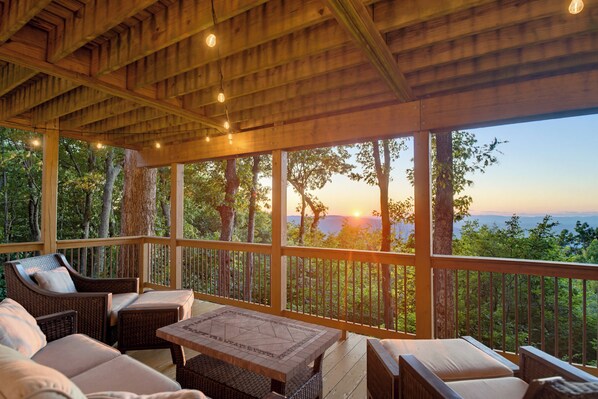 A covered outdoor seating area with wicker furniture and a view of the sunset over a forested landscape. - Sunset Views, Hot Tub, and Firepit in Wintergreen! (Wintergreen Resort)