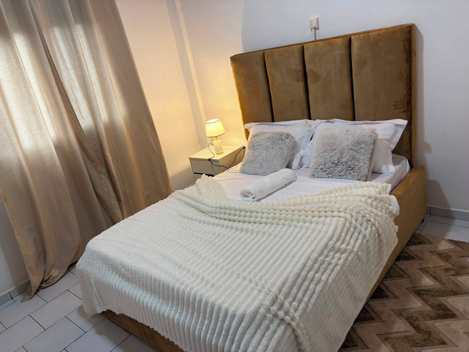 A neatly made bed with a white duvet and pillows, a wooden headboard, a bedside lamp, and a patterned floor.