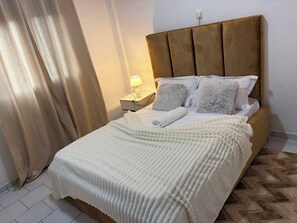 Una cama bien hecha con edredón blanco y almohadas, cabecera de madera, lámpara de noche y suelo estampado.