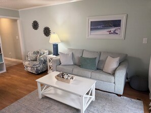 Ein Wohnzimmer mit grauem Sofa, einem gemusterten Sessel, einem weißen Couchtisch, einer Lampe und einem gerahmten Bild an der Wand.