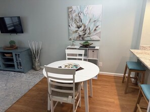 A dining area with a round table, white chairs, a flat-screen TV on the wall, and a painting above the table. - One Bedroom Beach Oasis in Ocean Creek Resort (Myrtle Beach)