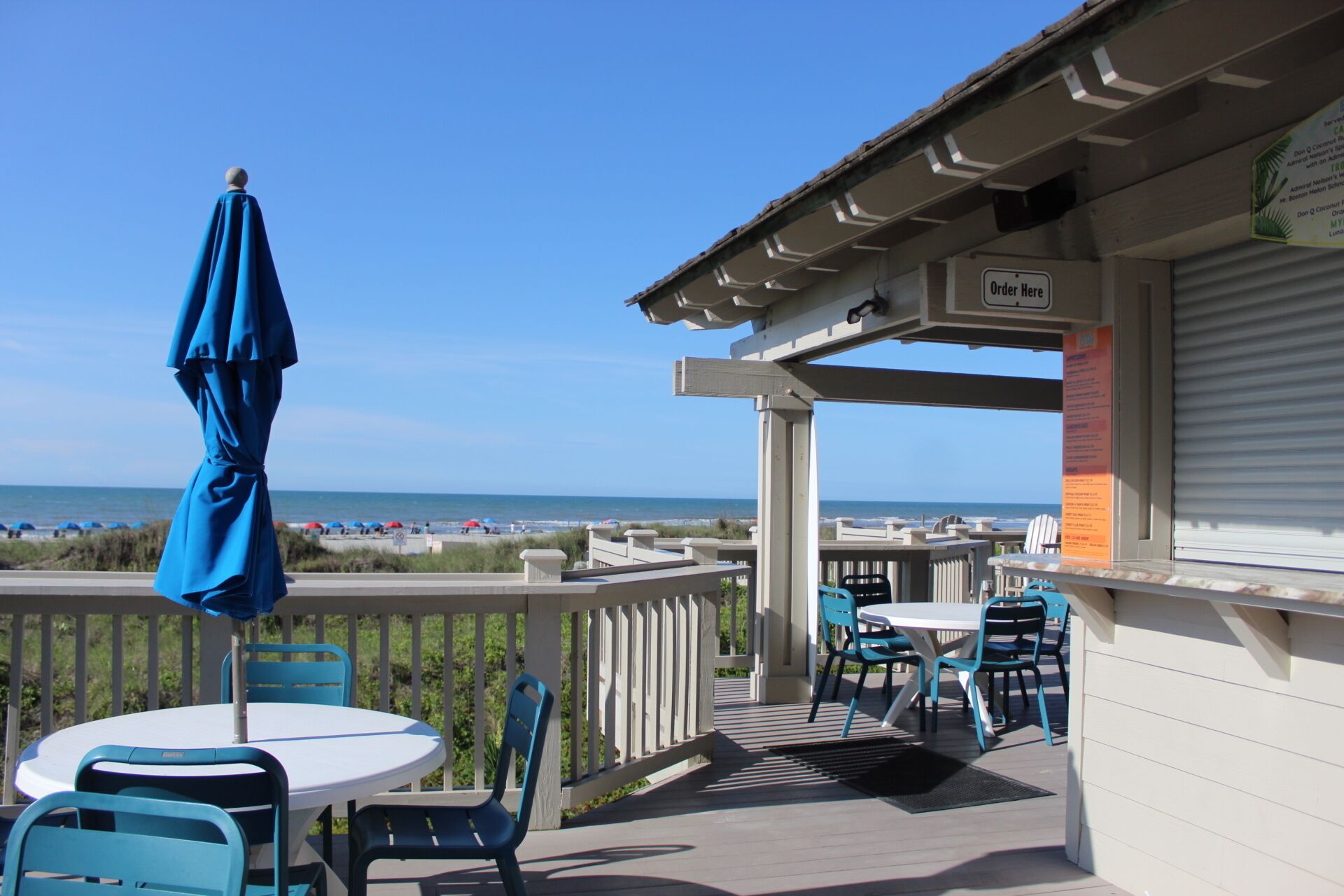 A covered outdoor seating area with a view of the beach, a table with chairs, and a sign indicating an order point.