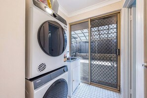 A laundry room with a front-loading washer and dryer, a small cabinet, and a sliding glass door leading to an outdoor area. - Mandurah Coastal Getaway|Dolphins|Beaches|Sleeps 6 (Mandurah)