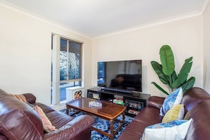 A living room with a television, leather sofas, a coffee table, and a large plant. - Mandurah Coastal Getaway|Dolphins|Beaches|Sleeps 6 (Mandurah)