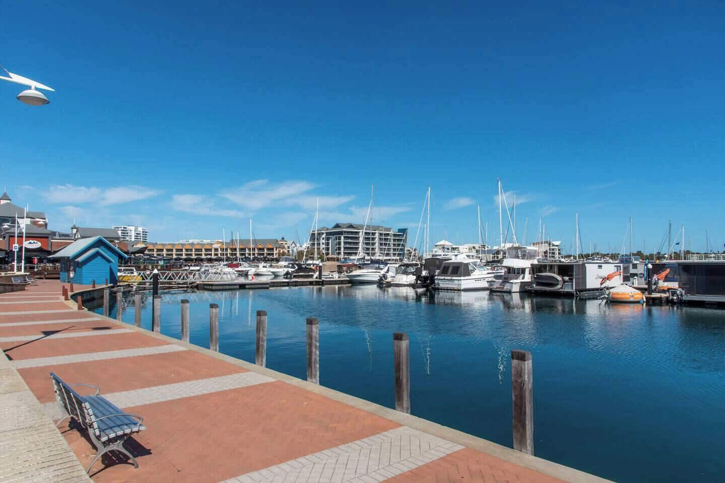 A marina with boats docked, a blue building, and a bench on a brick walkway.