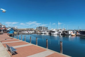 A marina with boats docked, a blue building, and a bench on a brick walkway. - Mandurah Coastal Getaway|Dolphins|Beaches|Sleeps 6 (Mandurah)