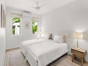 Ein Schlafzimmer mit zwei Betten, einem Deckenventilator, einem Fenster mit Blick ins Grüne und einem Nachttisch mit Lampe.