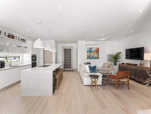 A modern kitchen with white cabinetry and a central island, a living area with a sofa and coffee table, and a flat-screen TV mounted on the wall. - Porters Place 2 | Modern West Coast Villa Near Holetown (Holetown)