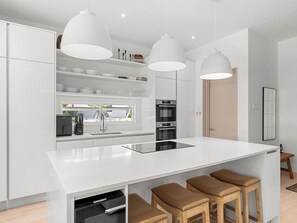 A modern kitchen with white cabinetry, a central island with stools, pendant lighting, and built-in appliances. - Porters Place 2 | Modern West Coast Villa Near Holetown (Holetown)