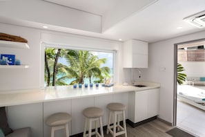 A modern kitchen with a large window, white cabinetry, and a bar area with stools. - Portobello Villa | Contemporary Beachfront Luxury on Barbados’ West Coast (St James)