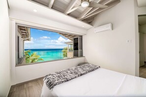 Una camera da letto con un'ampia finestra che si affaccia sull'oceano, un letto con lenzuola bianche e una ventola a soffitto.