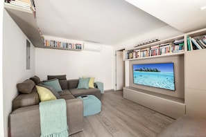 A modern living room with a grey sofa, a flat-screen TV, and bookshelves. - Portobello Villa | Contemporary Beachfront Luxury on Barbados’ West Coast (St James)