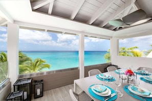 A dining area with a view of the ocean, a table set with blue and white tableware, and a ceiling fan. - Portobello Villa | Contemporary Beachfront Luxury on Barbados’ West Coast (St James)