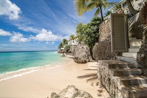 A beach with clear turquoise water, a sandy shore, and a building with a staircase leading to a door. - Portobello Villa | Contemporary Beachfront Luxury on Barbados’ West Coast (St James)