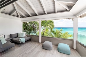 A modern interior with a grey sofa, a small round table, and a grey pouf. Large windows offer a view of the ocean and tropical foliage. - Portobello Villa | Contemporary Beachfront Luxury on Barbados’ West Coast (St James)