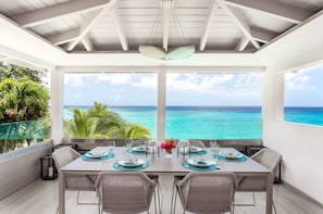 A dining area with a view of the ocean, featuring a table set for four with chairs, a ceiling fan, and a large window overlooking the sea. - Portobello Villa | Contemporary Beachfront Luxury on Barbados’ West Coast (St James)
