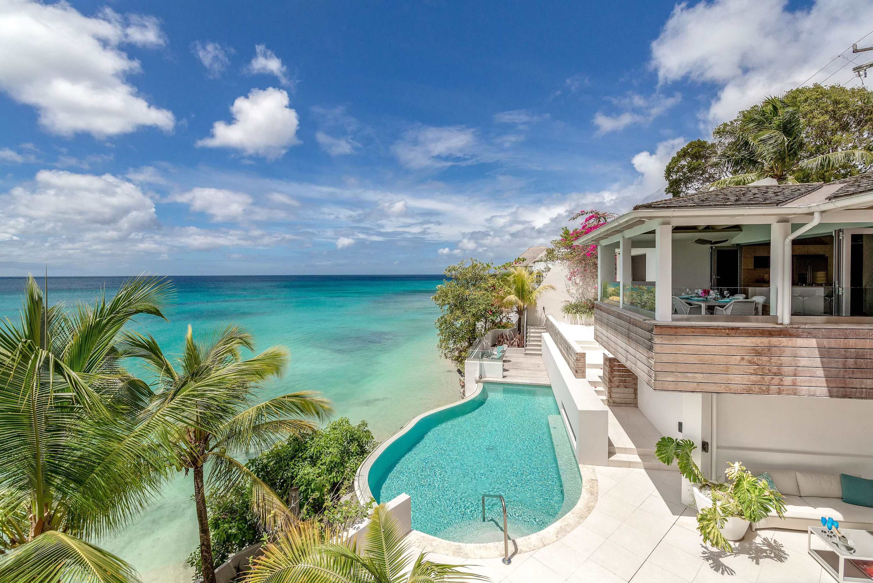 Um resort moderno com piscina e vista para um mar azul cristalino, rodeado por vegetação exuberante e uma área externa coberta.