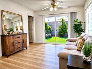 Ein geräumiges Zimmer mit einem Holzschrank, einem Sofa, einem Deckenventilator und Blick auf einen Garten mit Terrasse und Grünflächen.