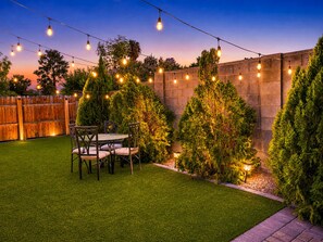 Patio trasero con cerco de madera, una mesa con sillas y luces colgantes.