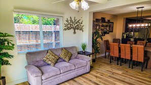 A living room with a sofa, a dining area with chairs, a ceiling fan, and a kitchen area. - Large Vacation House w/ Game Room 10-min Down Town (Las Vegas)