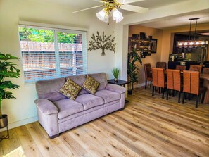 A living room with a sofa, a dining area with chairs, a ceiling fan, and a kitchen area. - Large Vacation House w/ Game Room 10-min Down Town (Las Vegas)