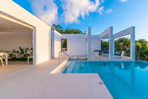 Ein moderner Poolbereich im Freien mit blauem Himmel, einer überdachten Lounge mit einer Couch und Sesseln sowie einem Weg zum Pool.
