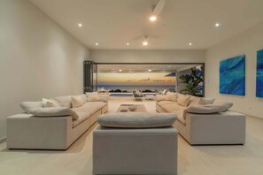 A modern living room with a large window overlooking a scenic view, a ceiling fan, and contemporary furniture. - Monkey Ridge | Contemporary Luxury with Panoramic Sea Views (Lower Carlton)