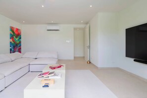 A modern living room with a sectional sofa, a coffee table with books and a board game, a flat-screen TV, and a colorful abstract painting on the wall. - Monkey Ridge | Contemporary Luxury with Panoramic Sea Views (Lower Carlton)