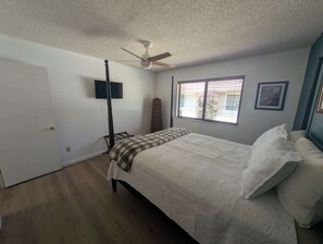 Ein Schlafzimmer mit einem Bett, einem Deckenventilator, einem Fenster mit Jalousien, einem Schminktisch und einem an der Wand befestigten Fernseher.