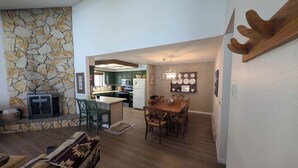 A spacious living area with a stone wall, a fireplace, a dining table with chairs, and a kitchen in the background. - Paw's For Awhile - 55 Ponderosa (Plumas Eureka)
