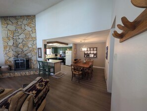 A spacious living area with a stone wall, a fireplace, a dining table with chairs, and a kitchen in the background. - Paw's For Awhile - 55 Ponderosa (Plumas Eureka)