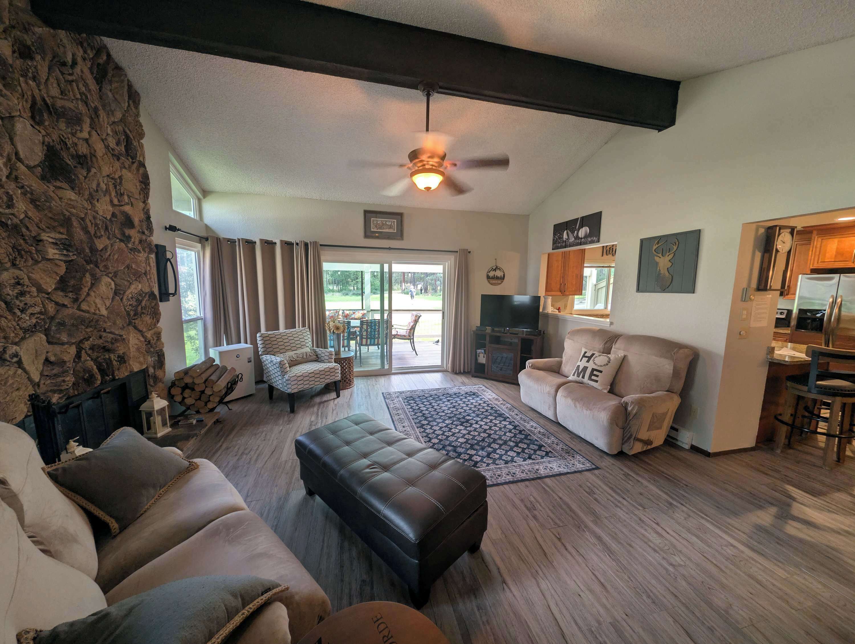 Una sala de estar con pared de piedra, una chimenea, un ventilador de techo, un sofá, una mesa de centro y una zona de cocina con un refrigerador.