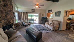 A living room with a stone wall, a fireplace, a ceiling fan, a sofa, a coffee table, and a kitchen area with a refrigerator. - Pondy - 101 Ponderosa (Plumas Eureka)