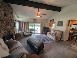 A living room with a stone wall, a fireplace, a ceiling fan, a sofa, a coffee table, and a kitchen area with a refrigerator. - Pondy - 101 Ponderosa (Plumas Eureka)