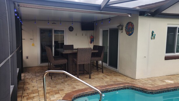 A covered outdoor area with a pool, a dining table with chairs, and a television mounted on the wall. - 3-bedroom house in peaceful Port Richey with AC comfort (Port Richey)