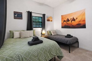 A bedroom with two beds, a window with curtains, a lamp, and a desert-themed painting.