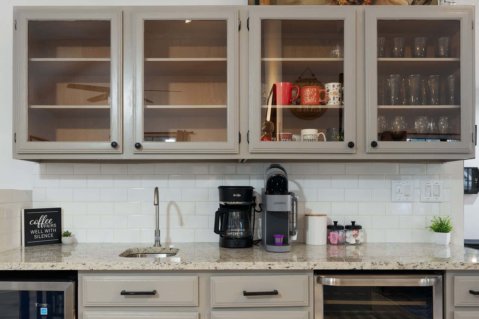 A kitchen with a coffee maker, a sink, and cabinets with glass doors containing mugs and glasses.