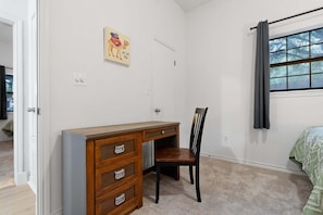 A bedroom with a wooden dresser, a chair, a bed with a green bedspread, a window with curtains, and a wall-mounted picture.