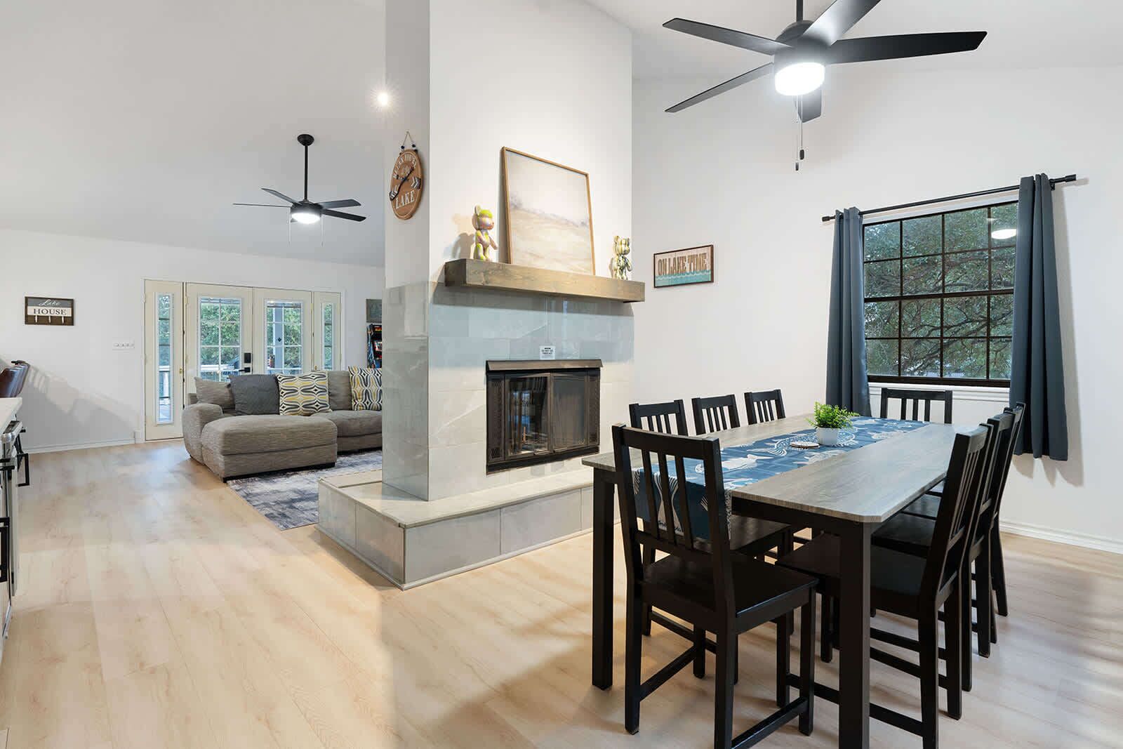 A modern living room with a fireplace, dining area with a table and chairs, and a ceiling fan.