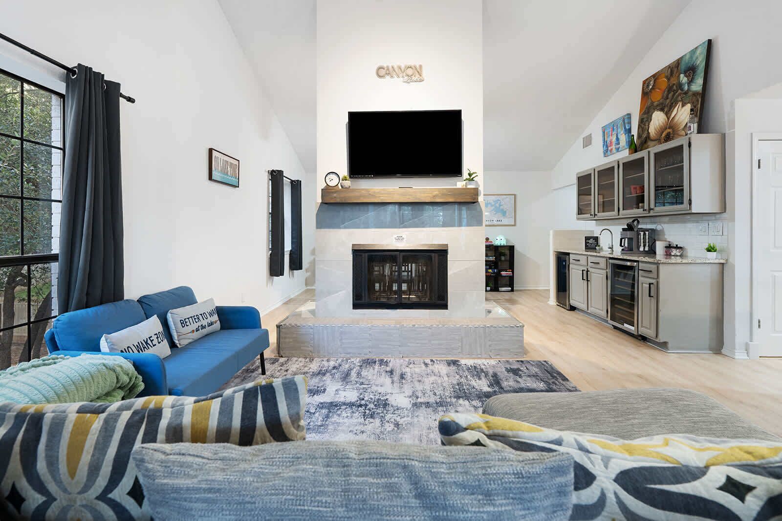 A modern living room with a fireplace, a flat-screen TV, a blue sofa, and a kitchen area with cabinets and appliances.