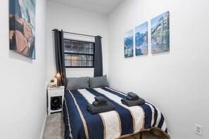 A bedroom with a bed, a window with curtains, a bedside table with a lamp, and three framed pictures on the wall.